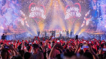 Un grupo de músicos sobre un escenario con pantallas iluminadas y confeti, frente a un público que sostiene teléfonos móviles