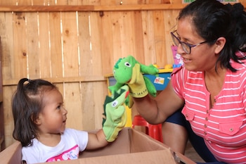 Una mujer adulta con lentes y una niña juegan con dos títeres de dedo de cocodrilos verdes. La niña está sentada en una caja de cartón, mirando a los títeres