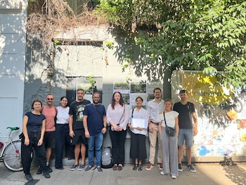 Diez personas, hombres y mujeres, posan al aire libre frente a una pared gris con vegetación y carteles sobre espacios verdes. Una mujer sostiene papeles
