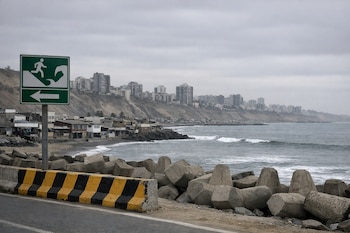 Una señal de evacuación de tsunami, barrera de carretera, bloques de cemento, playa, océano y edificios en un acantilado bajo cielo nublado.