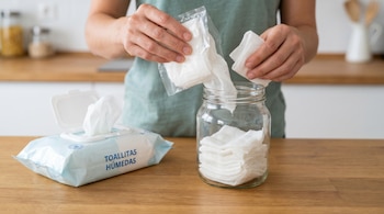 Manos transfiriendo toallitas húmedas de un paquete de recarga a un frasco de vidrio. Un paquete de toallitas abierto está a la izquierda sobre una encimera de madera.
