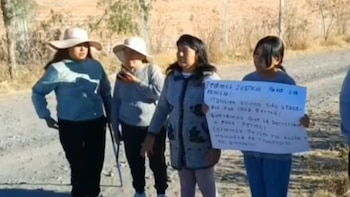 Cuatro mujeres en un camino de tierra, dos con sombreros, una sostiene una pancarta blanca con texto negro en español. Vegetación seca al fondo