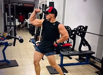 Un hombre joven de espaldas al equipo de un gimnasio, posa con el brazo flexionado mostrando su bíceps. Viste gorra, camiseta negra y shorts