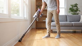 Un hombre utiliza una aspiradora sin cable de color gris y morado con un cepillo suave para limpiar el zócalo blanco de una pared en una habitación con piso de madera.