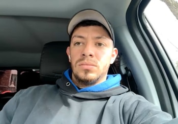Primer plano de Brayan López dentro de un vehículo, mirando al frente con expresión seria, lleva gorra, sudadera gris con azul y un auricular blanco en su oído izquierdo