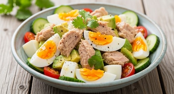 Un bol azul con ensalada de atún, trozos de aguacate, huevos cocidos cortados por la mitad, rodajas de pepino y tomates cherry sobre una mesa de madera.