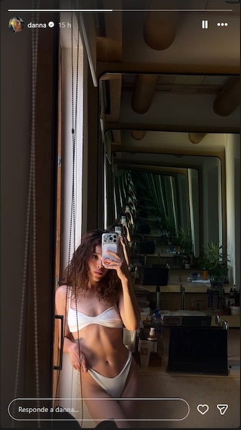 Mujer con cabello rizado y traje de baño blanco se toma una selfie con su teléfono frente a un espejo. Se observa un interior