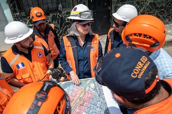 Un grupo de personas con chalecos naranjas, cascos de seguridad y gorras observa un mapa extendido en una calle