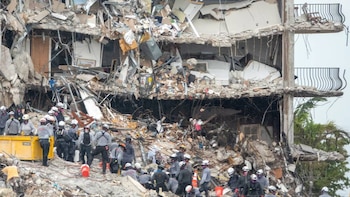 Restos de un edificio parcialmente derrumbado, con pisos y balcones expuestos. Rescatistas con cascos y máscaras trabajan entre los escombros