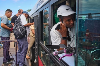 Varias personas suben a un vehículo improvisado en La Habana; un hombre con casco blanco y camiseta blanca mira desde una ventana, sosteniendo un teléfono