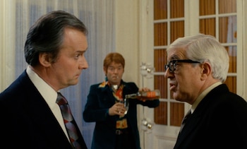 Primer plano de dos hombres de perfil, uno con cabello oscuro y corbata, otro con cabello blanco y gafas, conversando; un tercer hombre sirve una bebida al fondo