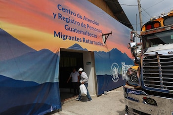 Funcionarios en uniforme azul atienden a personas frente a computadoras y sellos junto a un cartel del Instituto Guatemalteco de Migración