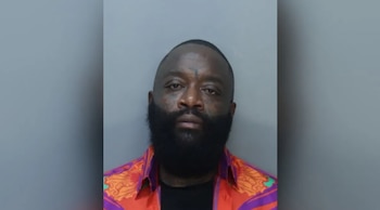Retrato frontal de un hombre con barba negra densa y camisa de estampados coloridos sobre un fondo gris