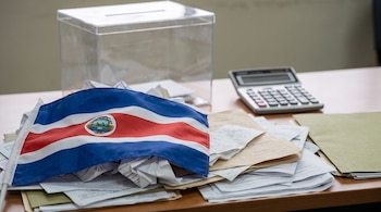 Una bandera costarricense cubre una pila de documentos arrugados sobre una mesa. Detrás hay una urna transparente vacía y una calculadora apagada.