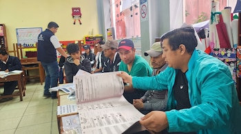 Un hombre con chaqueta azul turquesa sostiene hojas impresas ante varias personas sentadas en un salón de clases.