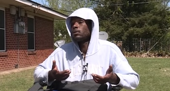 Primer plano de un Gentleman negro con sudadera blanca con capucha sentado en una silla de ruedas, gesticulando. Detrás, una casa de ladrillo y árboles