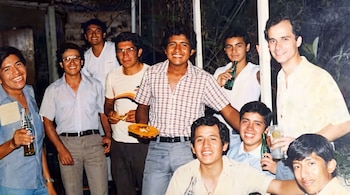 Nueve hombres jóvenes posan juntos; algunos sostienen botellas de vidrio, un vaso y un plato pequeño con comida