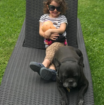 Una niña con gafas de sol y pelo rizado sentada en una tumbona negra, abrazando una muñeca, junto a un Bulldog negro acostado en el césped