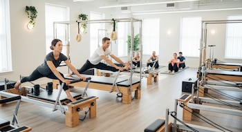 Un estudio de Pilates luminoso con seis personas realizando ejercicios en Reformers, Cadillacs y colchonetas.
