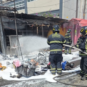 Tres bomberos con uniforme y casco rocían espuma con mangueras sobre una estructura calcinada y escombros en una calle