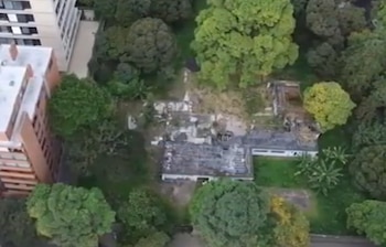 Suelo de concreto agrietado con pilas de ladrillos, restos de muros dañados, árboles al fondo y una persona a la distancia