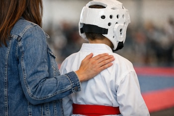 Una madre con chaqueta de mezclilla apoya su mano en el hombro de su hijo, quien viste un uniforme de karate blanco, un cinturón rojo y un casco protector blanco.