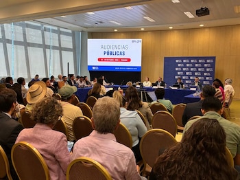 Cuatro personas sentadas ante una mesa con mantel azul, micrófonos, computadoras y un panel de fondo con logotipos institucionales