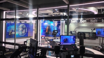 Un estudio de noticias de televisión vacío con pantallas encendidas, equipos de cámara, mesas de control con consolas y monitores, y reflectores de iluminación.