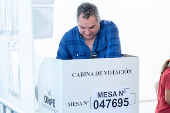 Un hombre de cabello canoso y camisa a cuadros azul, Rafael Belaúnde, vota dentro de una cabina de votación blanca con el texto "CABINA DE VOTACIÓN" y "ONPE"