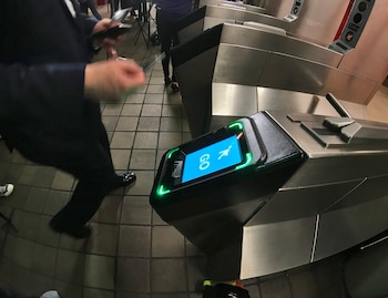 Una persona se aproxima a un torniquete del metro con pantalla azul que muestra “GO” y luces verdes. Otros torniquetes de metal se ven al fondo