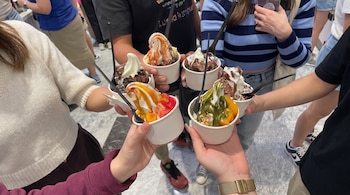 Primer plano de seis manos de varias personas que sostienen copas de helado con toppings de frutas, chocolate, salsa y granola.