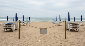 Playa de arena, mar, cielo nublado, sombrillas azules plegadas, reposeras blancas apiladas, dos postes con una cuerda, cartel rectangular en blanco.