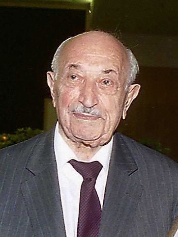 Un hombre mayor con bigote y poco cabello viste un traje oscuro con rayas diplomáticas, camisa blanca y corbata