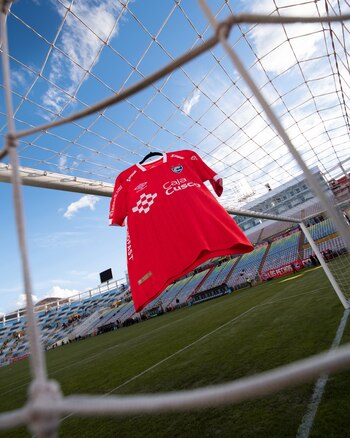 Una camiseta roja de fútbol colgada en la red de un arco dentro de un estadio con tribunas de colores y césped verde