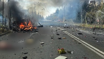 Un vehículo calcinado en llamas en el borde de un camino asfaltado cubierto de escombros, cables y humo