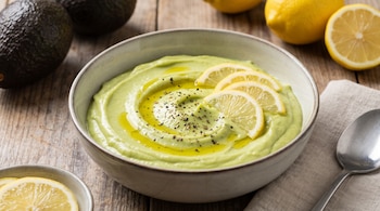 Cuenco con crema verde decorada con rodajas de limón y pimienta, junto a paltas enteras, limones y una cuchara sobre una mesa de madera.