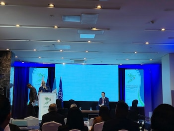 Una vista de una sala de conferencias con un hombre hablando en un podio frente a una pantalla grande azul claro y luces de techo empotradas