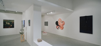 Interior de una galería de arte con paredes blancas y suelo gris, mostrando varias obras: una casa negra en patas, un cuadro texturizado, dos lienzos gráficos, y una escultura suspendida