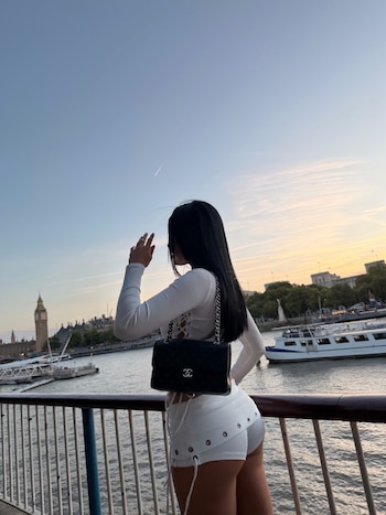 Una mujer de cabello negro y ropa blanca posa junto a una barandilla frente al río Támesis, con la torre del Big Ben al fondo