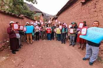 Un grupo de personas sostiene carpetas rojas y carteles de bienvenida en una calle de tierra flanqueada por muros de adobe