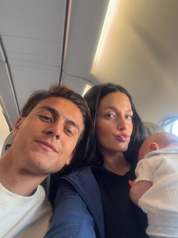 Primer plano de Paulo Dybala, Oriana Sabatini con su hija en brazos, dentro de la cabina de un avión. Dybala sonríe y Sabatini hace boca de pato