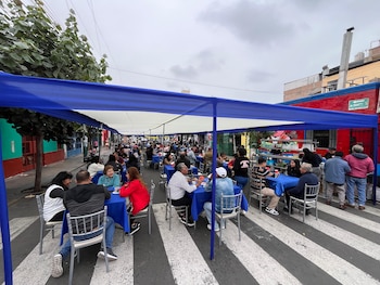 Multitud de personas sentadas en mesas con manteles azules bajo carpas blancas y azules en una calle con paso de cebra. Varios edificios coloridos en el fondo