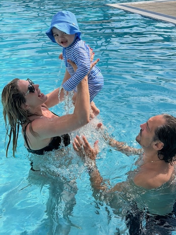 Una mujer con gafas oscuras sostiene en alto a un bebé con traje a rayas azules y gorro, junto a un hombre dentro de una piscina