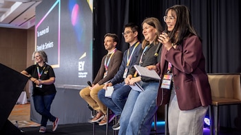 Cuatro jóvenes sentados sobre un escenario con micrófonos y libretas frente a una pantalla con el logotipo de EY