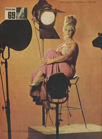 Nelida Lobato, rubia y con tiara, sentada de perfil en un taburete alto, viste un vestido rosa con lentejuelas y tacones plateados, rodeada de focos de estudio