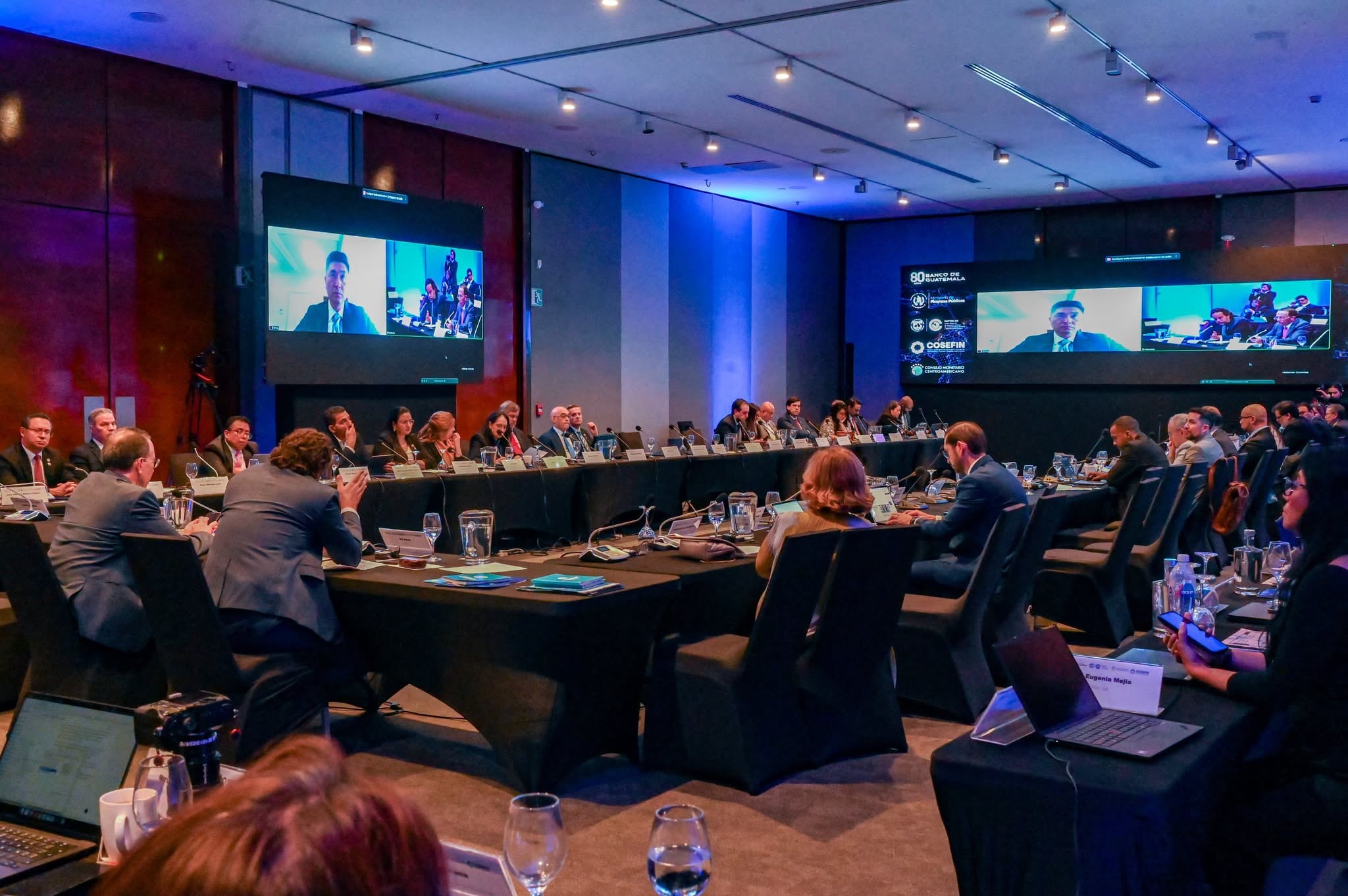 Participantes de una conferencia de alto nivel asisten a una sesión híbrida con intervenciones presenciales y virtuales proyectadas en pantallas, incluyendo logos del Banco de Guatemala y COSEFIN. (Banco de Guatemala)