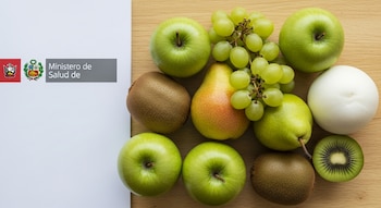 Manzanas verdes, uvas verdes, peras, kiwis, uno de ellos cortado, y una fruta blanca redonda sobre una mesa de madera, con un folleto del Ministerio de Salud.