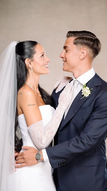 Mujer de novia con velo, vestido blanco, guantes de encaje y collar. Hombre de traje oscuro, camisa blanca, corbata y flor. Ambos se abrazan y sonríen