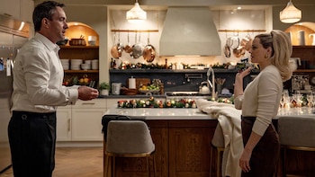 Matthew Macfadyen, de pie con camisa blanca, frente a una mujer rubia con suéter y falda que sostiene un camión de juguete en una cocina decorada