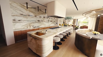 Cocina de concepto abierto con isla central de mármol, fregadero y fogones. Muebles de madera, estantes flotantes, taburetes. Salón y comedor al fondo
